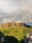 A hill with green grass towards it and cloudy sky above it. Therecrowns in the foreground.